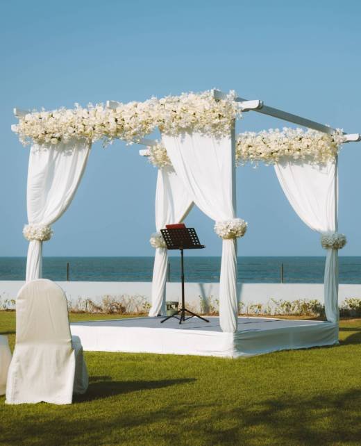 A wedding pavilion adorned by white curtains and artificial flowers set for an outdoor garden wedding by the sea